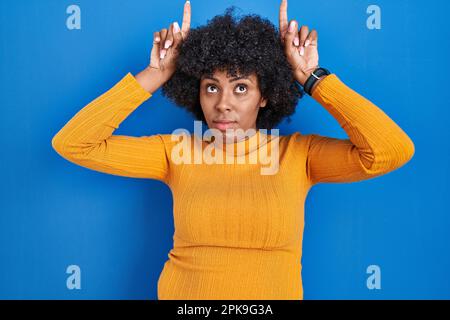 Schwarze Frau mit lockigem Haar, die auf blauem Hintergrund steht und eine lustige Geste macht, mit dem Finger über dem Kopf wie Bullenhörner Stockfoto