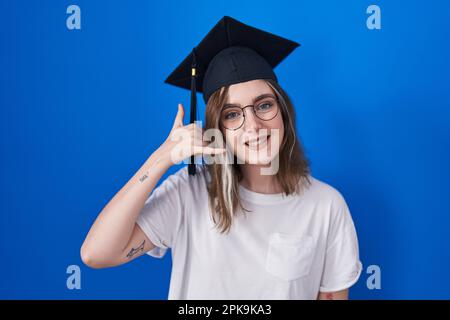 Blonde weiße Frau, die eine Abschlussmütze trägt und lächelt und Handygesten mit Hand und Fingern macht, als würde sie am Telefon reden. Kommunizieren von Konzepten. Stockfoto