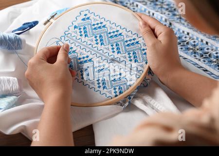 Eine Frau, die ein weißes Hemd mit blauem Faden am Holztisch stickt, Nahaufnahme. Ukrainische Nationalkleidung Stockfoto