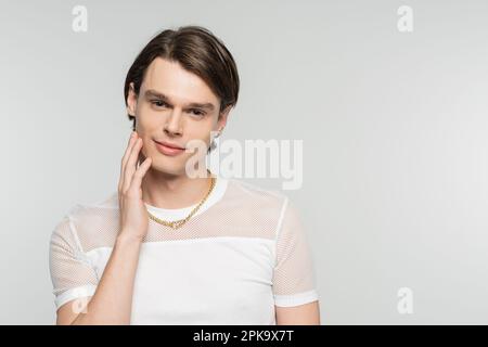 Positiver Pangender in weißem T-Shirt und goldener Halskette berührt Gesicht und lächelt vor der Kamera isoliert auf grauem Stockbild Stockfoto