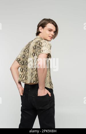 Trendige Pangender-Person in Bluse mit Tiermuster, die Hände in den Gesäßtaschen hält und vor der Kamera lächelt, isoliert auf grauem Stockbild Stockfoto