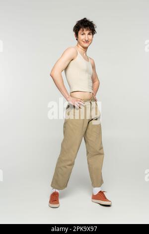 Die ganze Länge des fröhlichen Bigender-Modells in beiger Hose posiert mit Händen an der Hüfte auf grauem Hintergrund, Stockbild Stockfoto