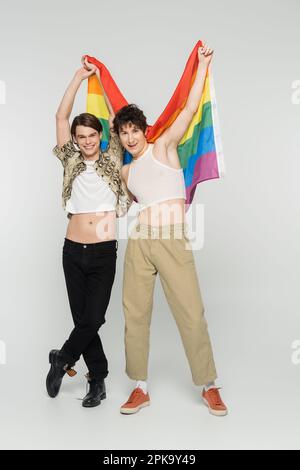 Volle Länge voller fröhlicher, großer Partner mit lgbt-Flagge in erhobenen Händen auf grauem Hintergrund, Stockbild Stockfoto