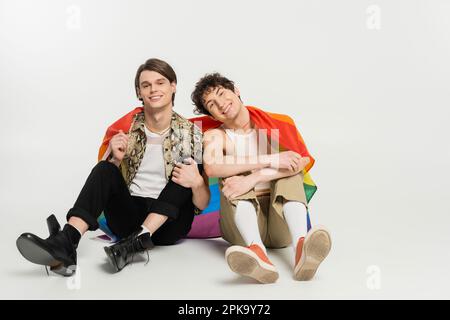 Die ganze Länge voller trendiger und fröhlicher nicht-binärer Partner mit Regenbogenflagge auf grauem Hintergrund, Stockbild Stockfoto