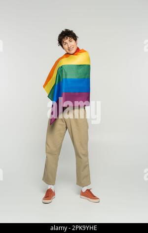 Die ganze Länge einer lächelnden, großen Person in beigen Hosen mit Regenbogenflagge auf grauem Hintergrund, Stockbild Stockfoto