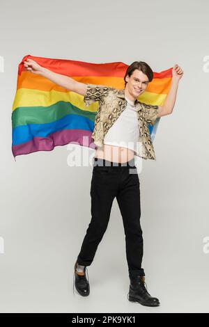 Eine elegante und sorgenfreie, nicht binäre Person in voller Länge, die mit Regenbogenfahne auf grauem Hintergrund läuft, Stockbild Stockfoto