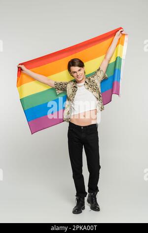 Durchgehende, sorgenfreie und trendige Person mit Regenbogenflagge in erhobenen Händen auf grauem Hintergrund, Stockbild Stockfoto