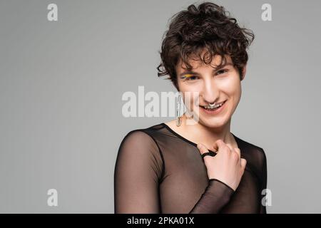 Glücklicher Bigender mit schwarzem, transparentem Oberteil und silbernem Zubehör, der auf die Kamera isoliert auf grauem, Stock Bild blickt Stockfoto