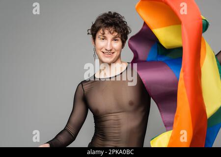 Fröhliche, nicht binäre Person mit schwarzem, transparentem Oberteil und silbernem Zubehör, das die lgbt-Flagge isoliert auf einem grauen Bild hält Stockfoto