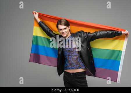 Fröhliche, große Person in schwarzer Lederjacke, die mit lgbt-Flagge posiert, isoliert auf grauem Stockbild Stockfoto