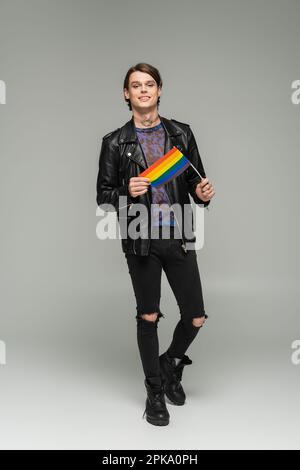 Durchgehende sorgenfreie Pangender-Person in schwarzer gerissener Hose und Lederjacke mit kleiner lgbt-Flagge auf grauem Hintergrund, Stockbild Stockfoto