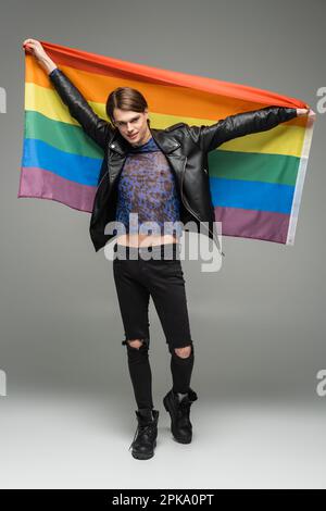 Die ganze Länge einer lächelnden Pangender-Person in einer schwarzen zerrissenen Hose und einer Lederjacke mit Regenbogenflagge auf grauem Hintergrund, Stockbild Stockfoto