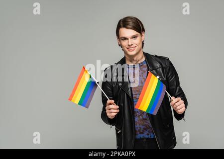 Fröhliche Pangender-Person in schwarzer Lederjacke, die kleine lgbt-Flaggen hält und vor der Kamera lächelt, isoliert auf grauem Stockbild Stockfoto