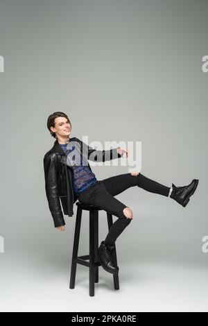 Sorgenfreier, großer Mensch in schwarz zerrissener Hose und Lederjacke, der auf einem hohen Hocker auf grauem Hintergrund posiert, Stockbild Stockfoto