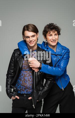 Glückliche, große Person in blauer Lederjacke, die einen stilvollen Partner umarmt, isoliert auf grauem, Stock Image Stockfoto