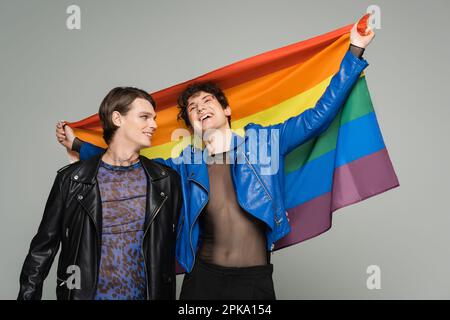 Sorgenfreier, wichtiger Mensch in blauer Lederjacke mit Regenbogenflagge in der Nähe eines trendigen Partners isoliert auf grauem Stockbild Stockfoto