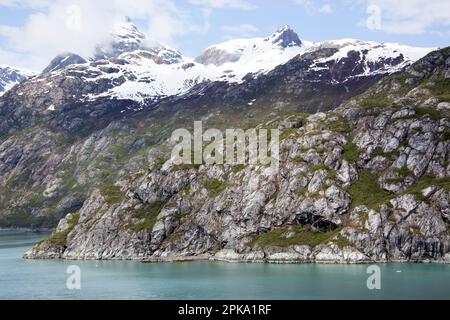 Der Glacier Bay-Nationalpark (Alaska) bietet im Frühsommer einen malerischen Blick auf eine felsige Küste und hohe schneebedeckte Berge. Stockfoto