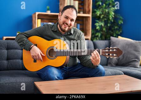 Junger hispanischer Mann spielt klassische Gitarre und sitzt zu Hause auf dem Bett Stockfoto