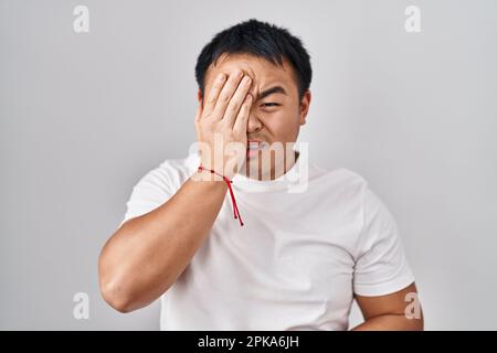 Ein junger chinesischer Mann steht über weißem Hintergrund und gähnt müde und bedeckt das halbe Gesicht, Auge und Mund mit der Hand. Das Gesicht tut weh. Stockfoto