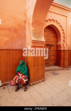 Marokko, Marrakesch, Madersa Ben Youssef Madrasa, schöne Architektur, sitzende Frau mit grünem Schleier vor dem braunen orangefarbenen Gebäude Stockfoto