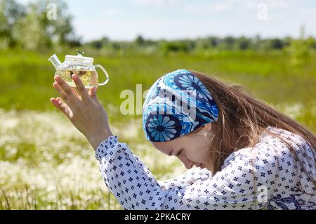 Frau mit transparenter Teekanne und Kräutertee. Sammelt frische grüne Kräuter, ernten und machen gesunden Tee. Verschwommenes Bild, selektiver Fokus Stockfoto