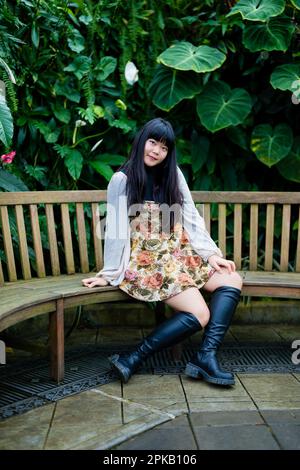 Junge asiatische Frau sitzt auf einer Teakbank in einem viktorianischen Garten | Z-Bein | Lächeln | warme Farbtöne | kniehohe Stiefel | kurzes Kleid Stockfoto
