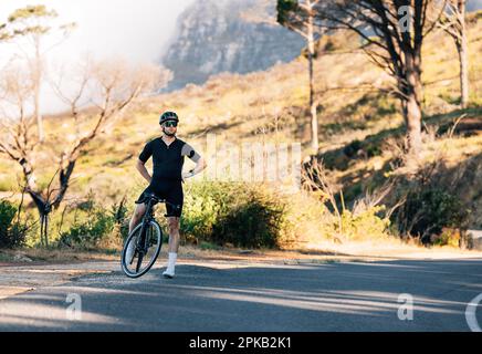 Professioneller Radfahrer in schwarzer Sportbekleidung, der sich am Straßenrand mit den Händen an der Hüfte entspannt Stockfoto