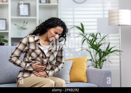 Kranke Erwachsene Frau, die allein zu Hause auf dem Sofa sitzt, starke Bauchschmerzen hat, lateinamerikanische Frau mit lockigem Haar im Wohnzimmer, die Hände an der Seite des Magens hält. Stockfoto