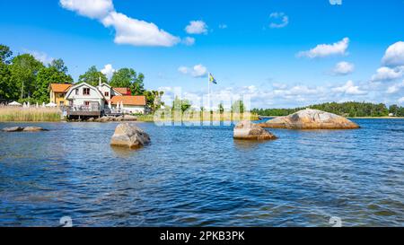 Sonniger Tag in einer kleinen Dorfbucht auf der Insel Vaxholm. An der Küste befindet sich das Hembygdsgard Museum und eine ruhige Terrasse mit Sitzbereichen für Gäste. Stockholmer Archipel, Schweden Stockfoto