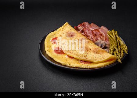 Köstliches frisches Omelett mit Kirschtomaten, Speck, Spargel und Gewürzen auf dunklem Betonhintergrund Stockfoto