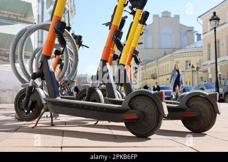 Elektrische Roller hintereinander auf dem Parkplatz auf Stadtgebäuden und im Hintergrund von Spaziergängern. Mietsystem, öffentliche E-Roller auf der Straße Stockfoto