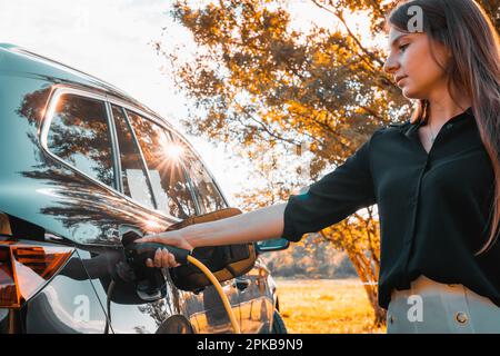 Weibliche Hand, die ein Kabelladegerät in ein Elektroauto steckt, beleuchtet von den Sonnenstrahlen, die durch die Zweige des grünen Baumes brechen, Nahaufnahme Stockfoto