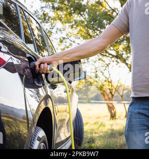 Junger Mann, der das Ladegerät in einem schwarzen Elektroauto anschließt, Konzept für erneuerbare Energien Stockfoto