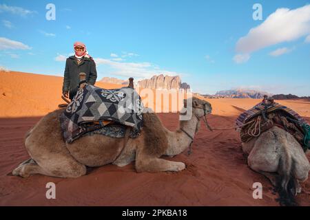 Wadi Rum, Jordanien - 19. Januar 2020: Unbekannter Mann in warmem Mantel und rot-weißer shemagh - traditioneller Beduinenkopfschal, Kamel auf roter Staubwüste Stockfoto