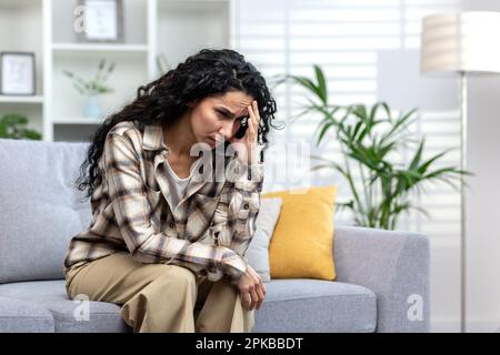 Einsame hispanische Frau, die allein auf dem Sofa im Wohnzimmer zu Hause sitzt, deprimierte Frau, die über schwierige Entscheidungen nachdenkt. Stockfoto