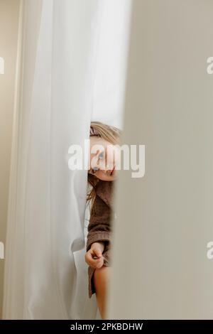 Ein Mädchen in einem braunen Bademantel sitzt auf der Fensterbank und guckt durch den Vorhang und lächelt. Stockfoto