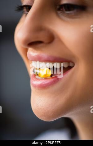 Multiethnische Happy Young Woman hält gelbe Pille im Mund zwischen den Zähnen in natürlichem Fischöl Omega 3 Vitaminpille steht zu Hause am Fenster Nahaufnahme Stockfoto