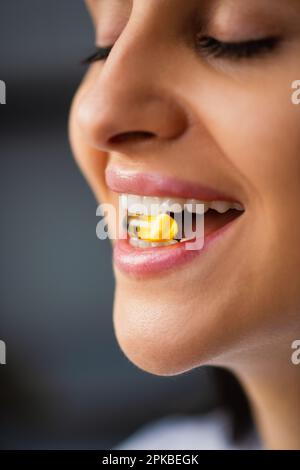 Multiethnische Happy Young Woman hält gelbe Pille im Mund zwischen den Zähnen in natürlichem Fischöl Omega 3 Vitaminpille steht zu Hause am Fenster Nahaufnahme Stockfoto