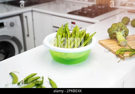 Moderne Küche mit grünem Gemüse, romano-Helda-Bohnen, grünen Erbsen und Brokkoli auf dem Tisch. Helle, weiße Küche. Der Begriff der Gesundheit Stockfoto
