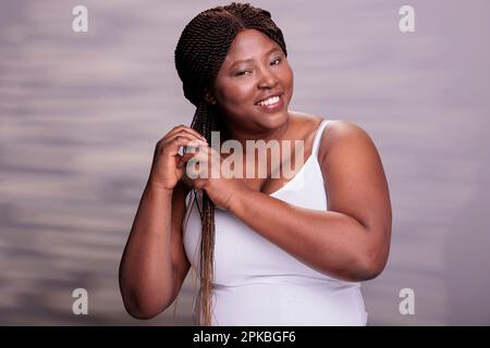 Körperpositive Frau stylen Haare und machen Haarpflegemortrait. Junger afroamerikanischer Kurvengeist, der in die Kamera schaut und im Studio Zöpfe für Haarpflegeprodukte herstellt Stockfoto