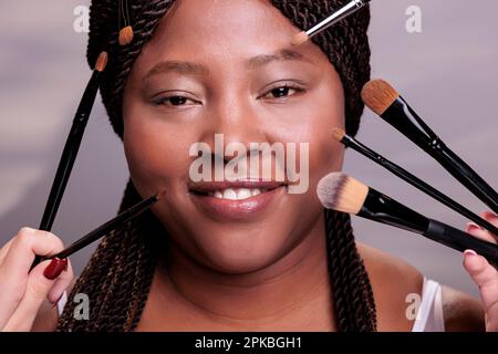 Eine afroamerikanische Frau, die ein professionelles Make-up-Porträt bekommt. Make-up-Künstler tragen Augenschatten und Puder auf das Gesicht des Modells auf, indem sie dekorative Kosmetikpinsel verwenden Stockfoto