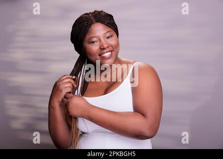 Körperpositive Frau, die langes braunes Haar mit Omelaseffekt geflochten hat. Eine ziemlich kurvige afroamerikanische Dame, die Zöpfe hält, in die Kamera schaut und fröhliche Emotionen im Studio ausdrückt Stockfoto