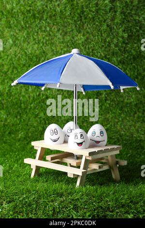 Eier mit fröhlichen Gesichtern, kleiner Picknicktisch und Sonnenschirm auf grünem Gras Stockfoto