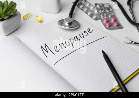 Notizbuch mit dem Wort Menopause, Stethoskop, Pillen und Zimmerpflanze auf weißem Hintergrund, Nahaufnahme Stockfoto