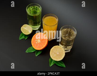 Drei Gläser mit Zitronensäften, Zitrone, Orangen und Limette, dekoriert mit Blättern auf schwarzem Hintergrund. Nahaufnahme. Stockfoto