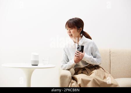 Frau sitzt auf dem Sofa mit Handy in der Hand und spricht mit AI-Lautsprecher Stockfoto