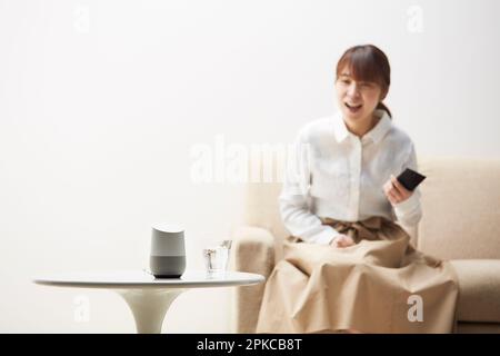 Frau sitzt auf einem Sofa mit einem Handy in der Hand und spricht mit einem AI-Lautsprecher Stockfoto