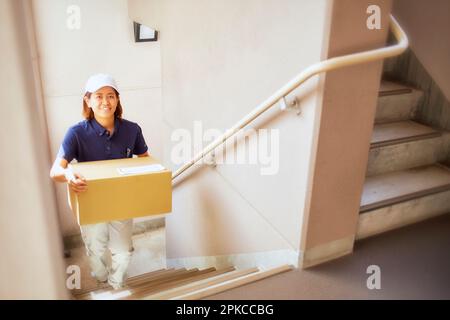 Eine Frau in Arbeitskleidung, die eine Pappschachtel die Treppe hoch trägt Stockfoto