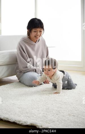 Ein Baby, das auf einem weißen Teppich krabbelt, während seine Mutter auf ihn aufpasst Stockfoto