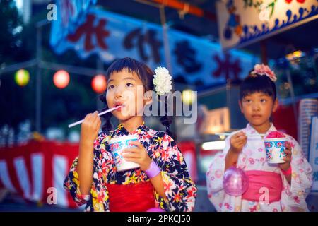 Ein Mädchen in Yukata isst rasiertes Eis, das an einem Festspielstand gekauft wurde Stockfoto